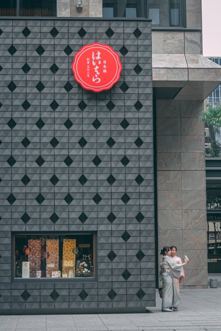 Two women in kimono beside a tiled shopfront with a red sign in Nihonbashi, Tokyo